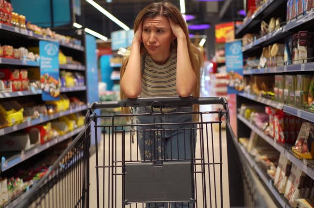 Young woman standing among the produce aisle at the supermarket and feeling worried about the increase in food prices
