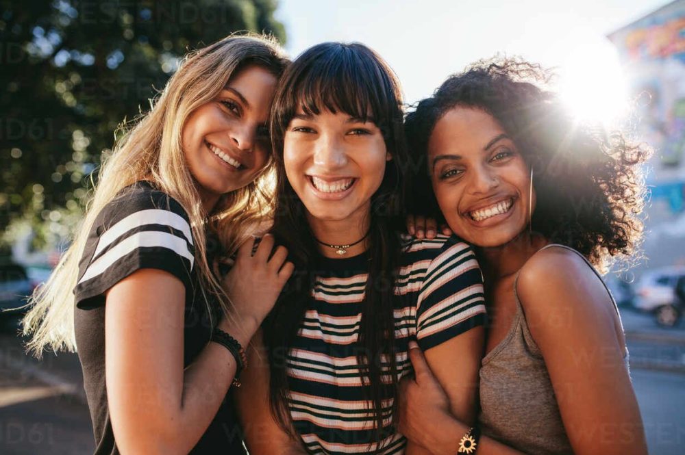 Three beautiful smiling girl friends standing together. Multi ethnic group of women outdoors in the city looking happy.